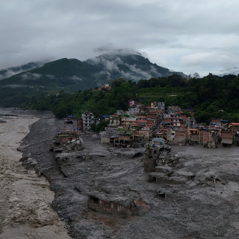 Aerial view of a river valley under low cloud. A broad grey flow of mud and rock has buried the flat ground beside a swollen brown river, leaving broken buildings half-submerged, with intact houses standing on the green slope above.