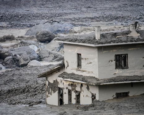 A two-storey house half-buried in grey mud and rock. Mud coats the roof, the walls and the window frames, and boulders and debris fill the ground around it.